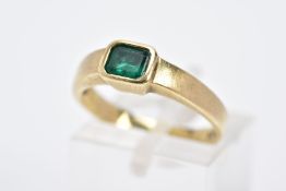 A YELLOW METAL EMERALD RING, the emerald cut within a collet mount to the plain polished band,