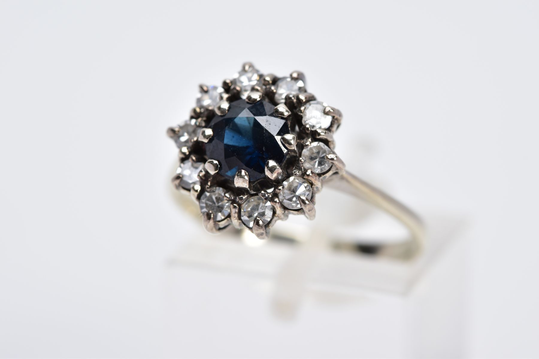 A WHITE METAL SAPPHIRE AND DIAMOND CLUSTER RING, designed with a central circular cut blue