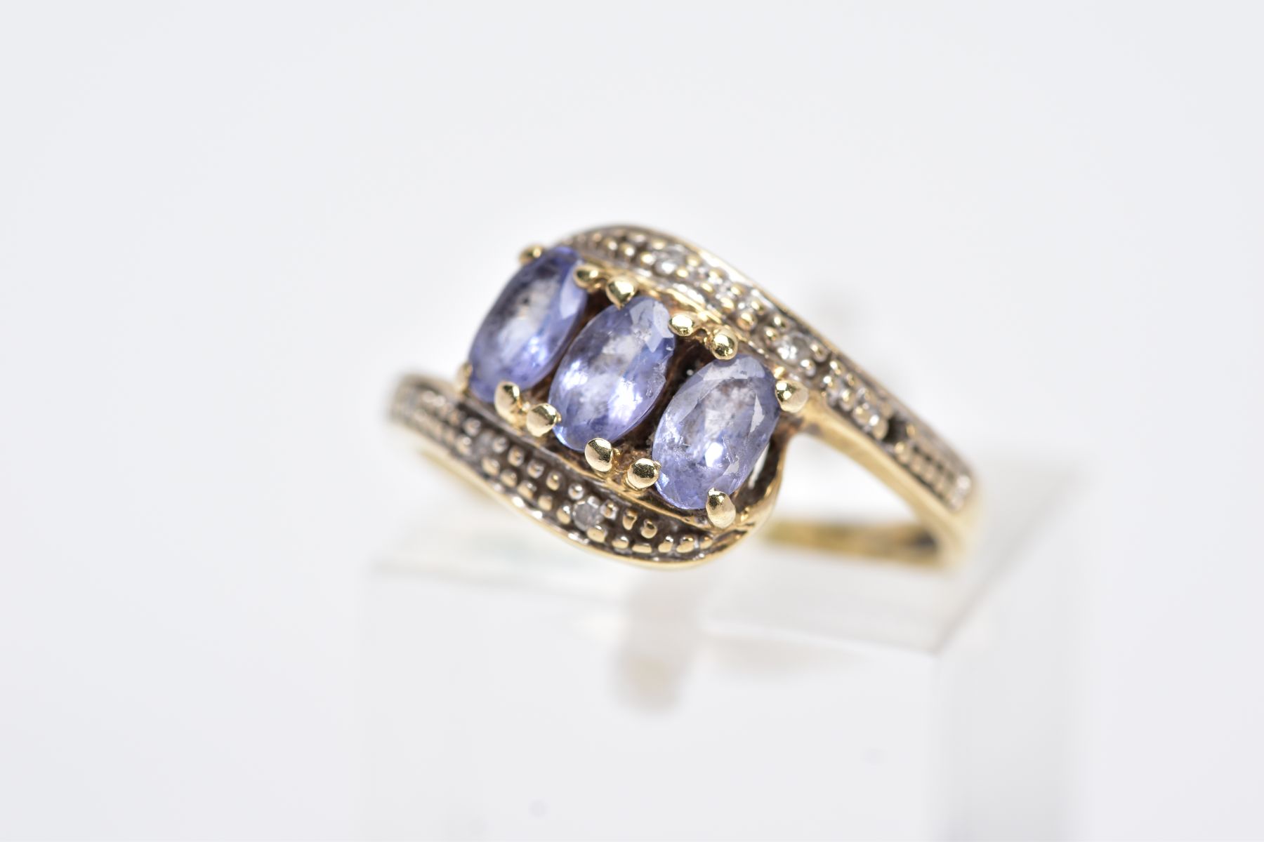 A YELLOW METAL GEM SET RING, of a crossover design, set with three oval cut purple stones assessed