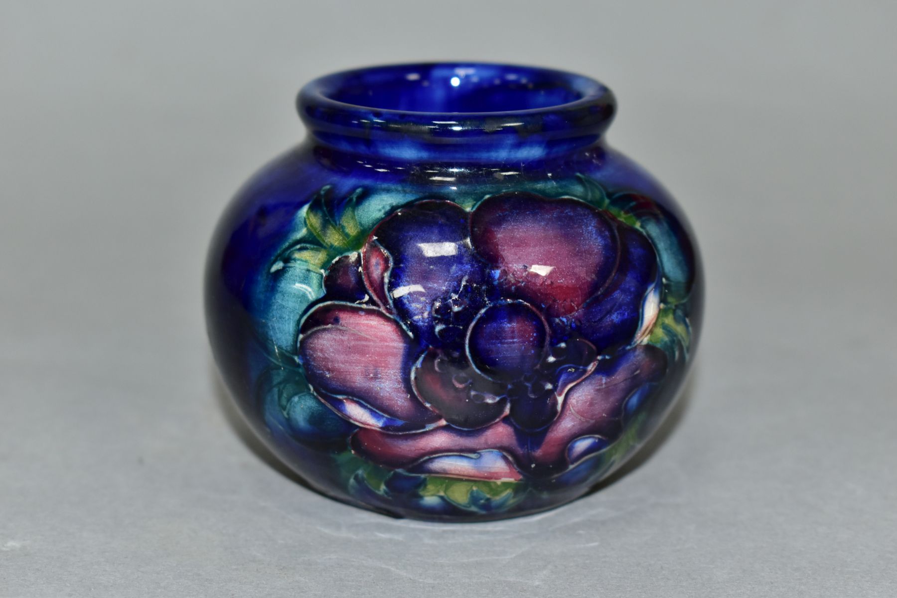 A MOORCROFT POTTERY SQUAT GLOBULAR VASE DECORATED WITH PURPLE/PINK ANEMONE, on a blue ground,