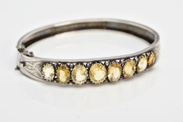 A WHITE METAL CITRINE HINGED BANGLE, designed with a row of graduated claw set, oval cut citrines,