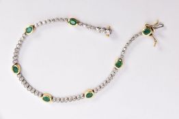 A MODERN 18CT GOLD DIAMOND AND EMERALD LINE BRACELET, seven oval mixed cut emeralds each measuring