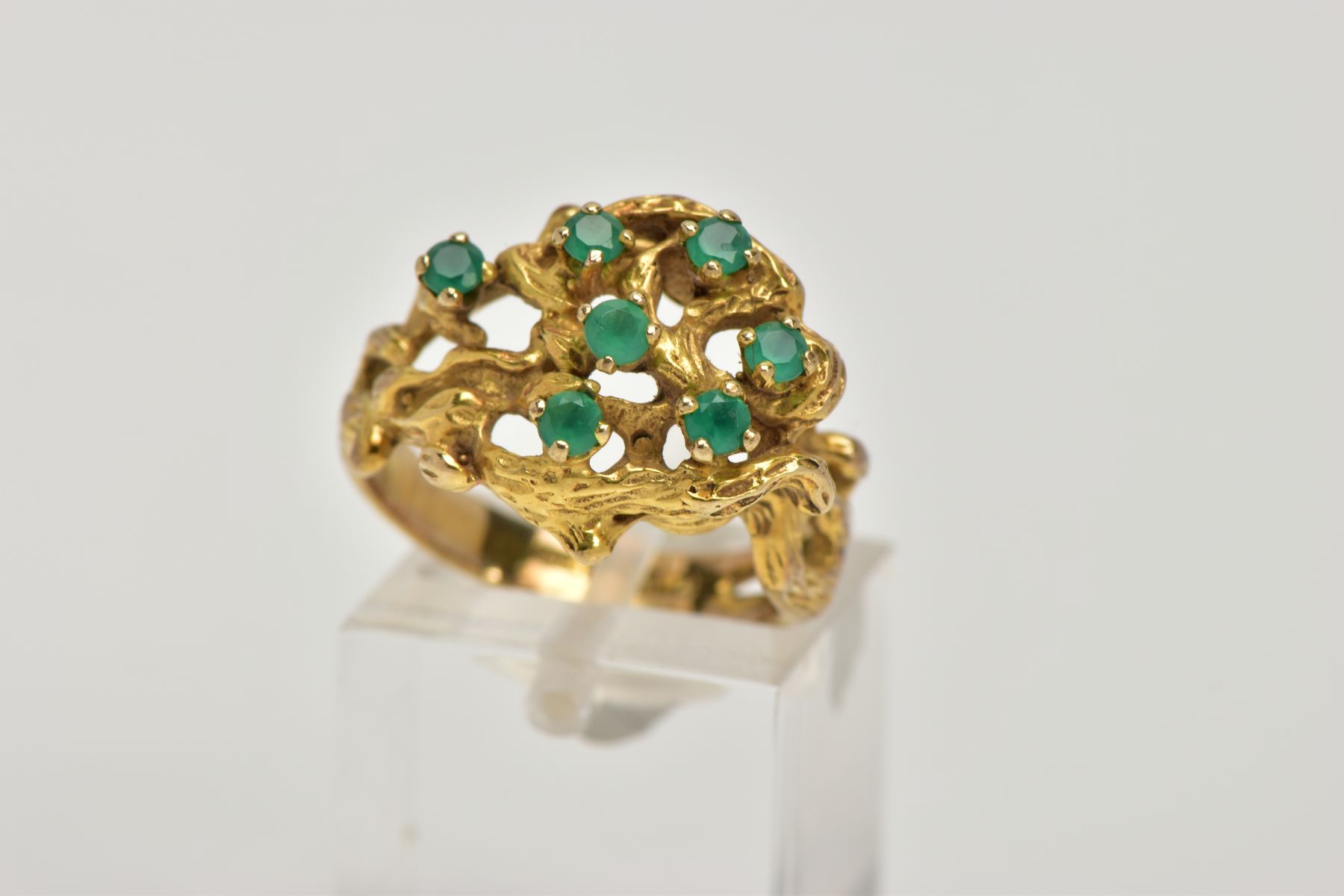 AN ABSTRACT GEM RING, of textured openwork design, claw set with seven circular cut green gems