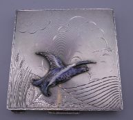 A square silver compact decorated with an enamelled game bird in a rustic landscape,