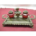 Ornate brass pen and ink stand with two cranberry glass inkwells with brass covers and a central
