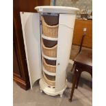 A tall cylindrical cream coloured cabinet, a pair of doors enclosing four tiers with wicker