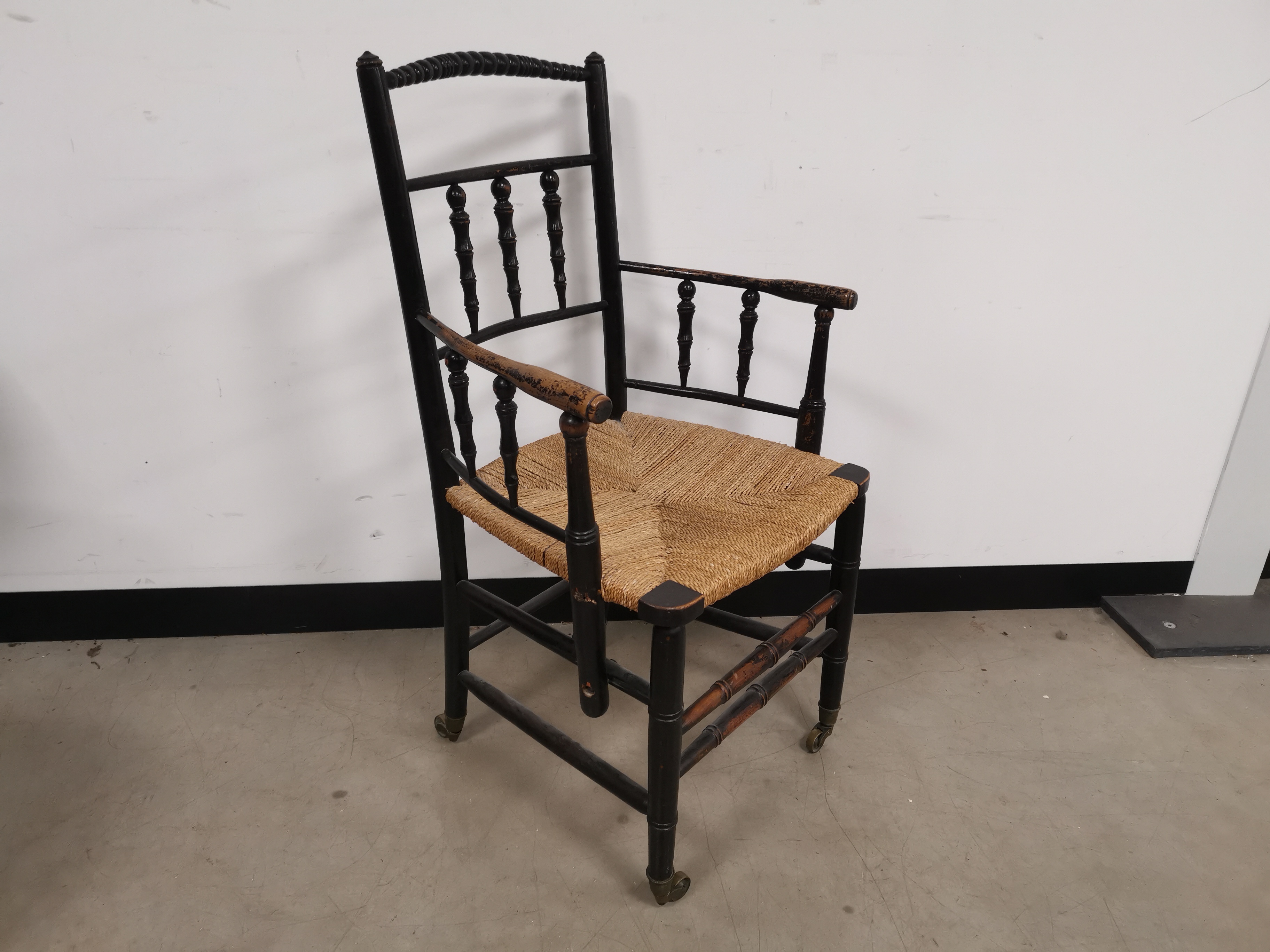 Black painted arm chair, with rush seat, turned spindle supports, set on brass castors. 57cm W x