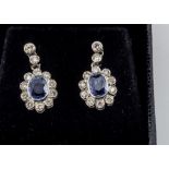 A pair of sapphire and diamond drop earrings, oval blue mixed cuts surrounded by a bezel of diamonds