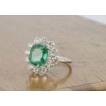 A contemporary emerald and diamond white metal cluster ring, the cushion cut emerald in claw setting