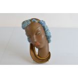 A Goldscheider pottery wall mask, modelled as a female face with blue hair, gilt scarf, red lips,