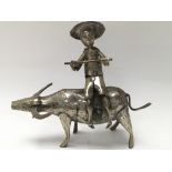 A white metal figure of an oriental man holding a stick whilst sat on a water buffalo.