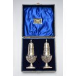 Cased Victorian pair of hallmarked silver peppers with repoussé decoration raised on square bases,