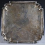 George V hallmarked silver salver of square form with shaped corners, raised on four hoof feet,
