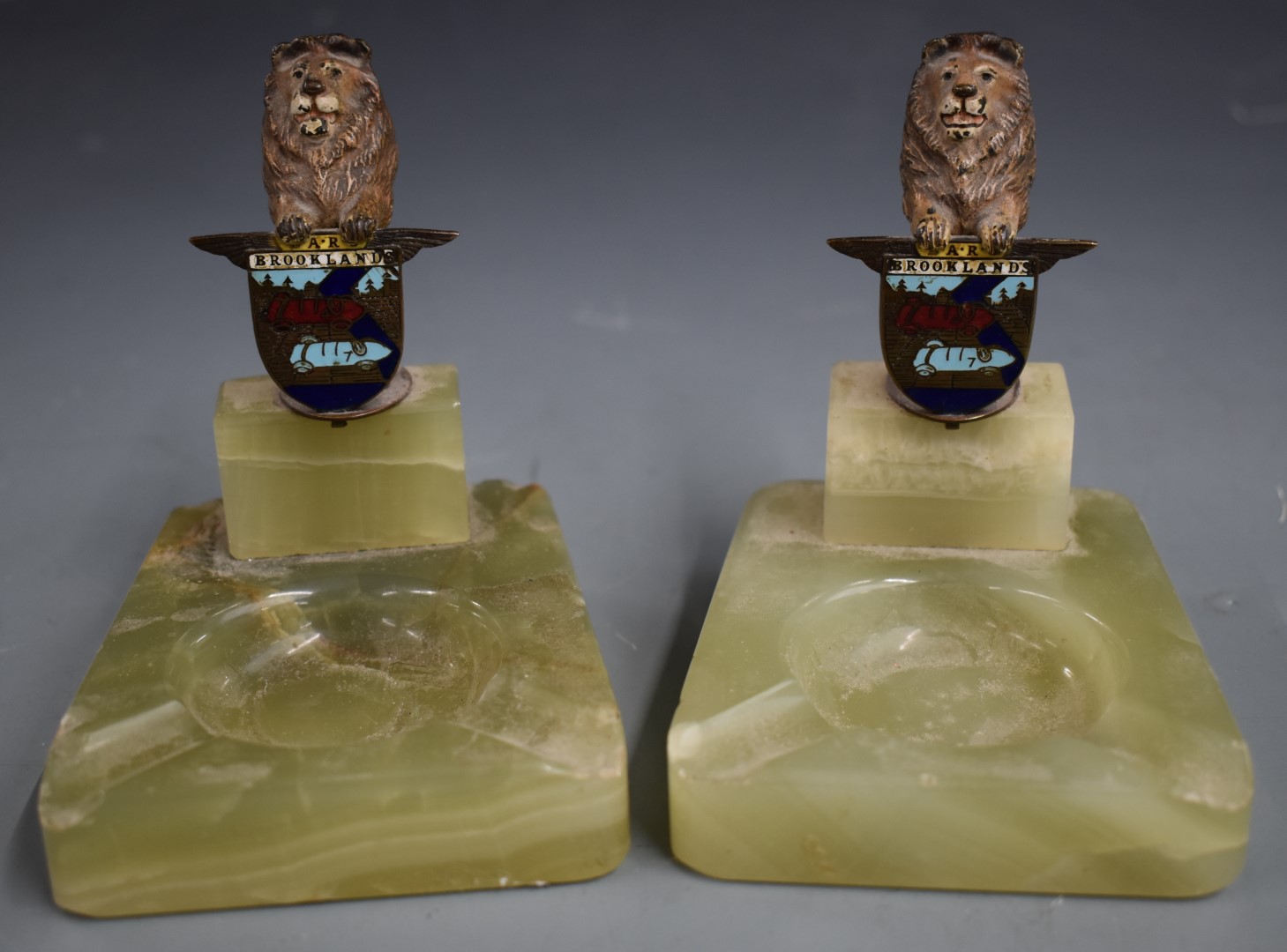 Pair of BARC Brooklands ashtrays, surmounted by cold painted bronze or similar lions holding