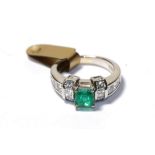 An emerald and diamond ring, the emerald-cut emerald in a white four claw setting, flanked by a trio