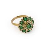 An emerald and diamond cluster ring, the central raised round cut emerald within a border of round