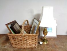 A wicker handled basket containing assorted framed pictures and prints, brass table lamp with shade.