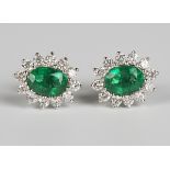 A pair of 18ct white gold, emerald and diamond earrings, each claw set with an oval cut emerald