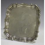 A George V silver card salver of square outline with piecrust rim, on four volute scroll feet,