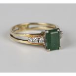 A gold ring, claw set with a rectangular cut emerald between circular cut diamond set three stone