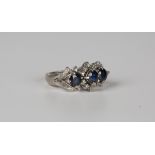 A white gold, sapphire and diamond ring, claw set with three cushion cut sapphires, otherwise set