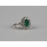 A platinum, emerald and diamond cluster ring, claw set with the oval emerald within a surround of