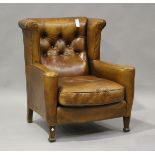 An early 20th century brown leather wingback armchair with buttoned back and scroll arms, raised