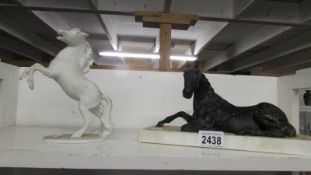 A black horse laying down on a white plinth and a white Kaiser porcelain rearing horse.
