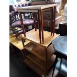 A teak nest of two tables and a light wood three tier trolley.