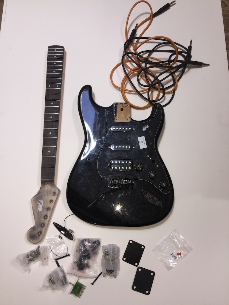 A black electric guitar in pieces. Stratocaster shape, pickguard has pots and pickups attached -