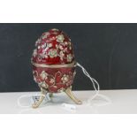 Russian style musical trinket box egg, raised on three feet, the red enamelled body set with paste