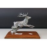 A white metal chrome car mascot in the form of a stag deer mounted on a wooden plinth.