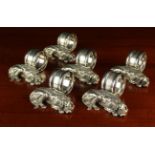 A Set of Six Ornamental Napkin Rings with white metal mounts cast in the form of recumbent