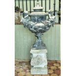 A weathered lead trophy shaped garden urn with removable lid, fruiting swags, seated cross legged