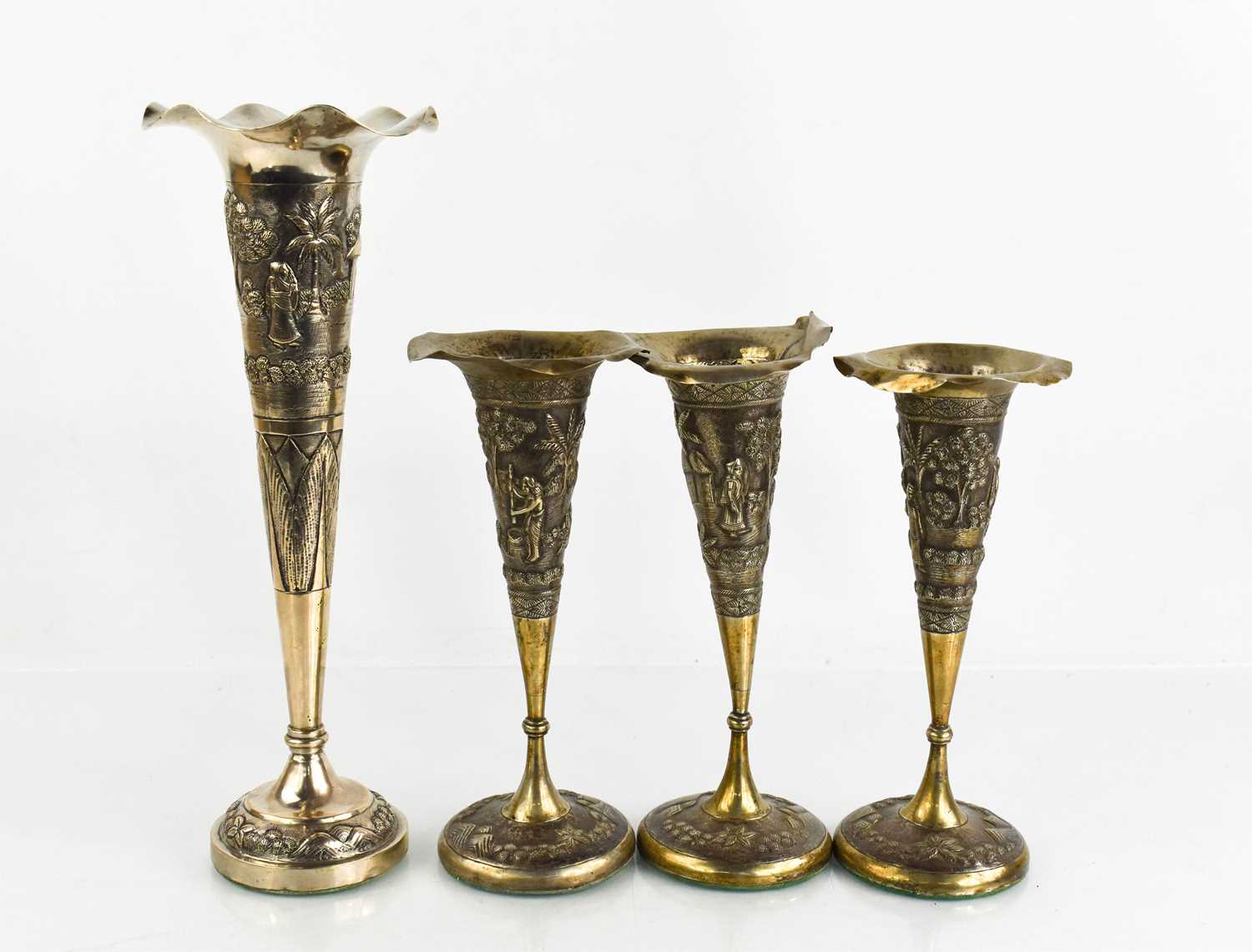 Four white metal Indonesian bud vases, depicting various domestic scenes, all with weighted bases.