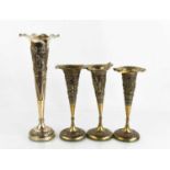 Four white metal Indonesian bud vases, depicting various domestic scenes, all with weighted bases.