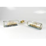 Two clear glass ships in bottles, one with four masts, set on naturalistic ground,