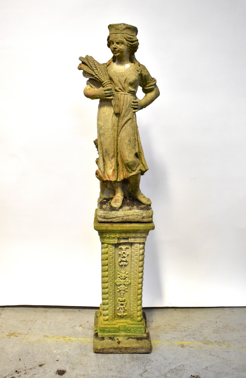 A composite stone garden statue in the form of a woman holding a wheatsheaf with one hand on her