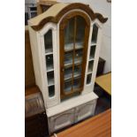 A white painted side cabinet, the arched glazed upper section above a base with two cupboard doors,