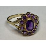 A 9 CARAT GOLD RING WITH AN AMETHYST SURROUNDED BY TEN SMALLER SIZE R/S - SMALL CHIP TO CENTRE STONE