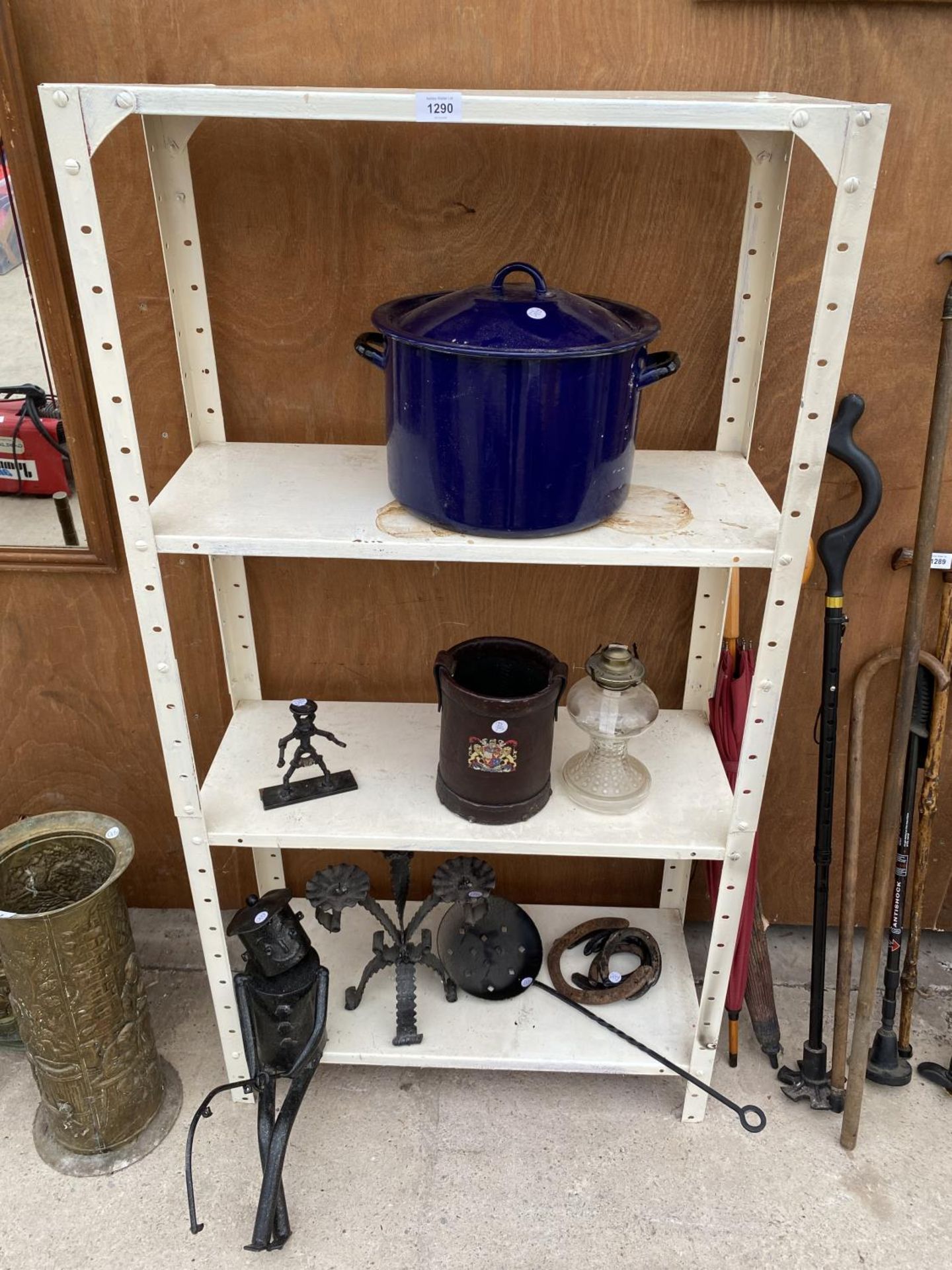 A FOUR TIER METAL SHELVING UNIT WITH CONTENTS TO INCLUDE A COOKING POT, HORSE SHOES AND AN OIL