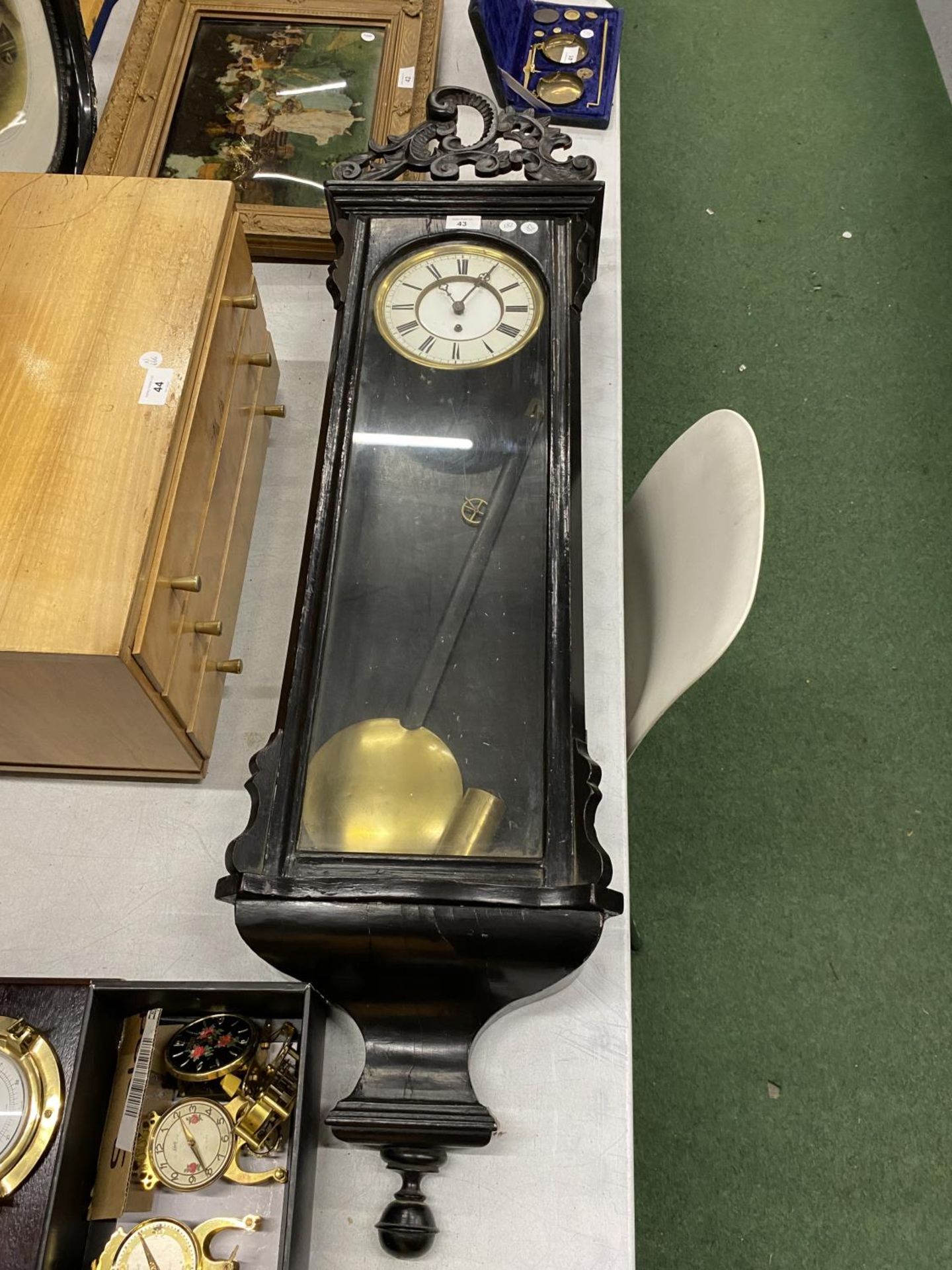 A LARGE WALL CLOCK IN EBONISED CASE WITH CARVED TOP WITH PENDULUM AND WEIGHT