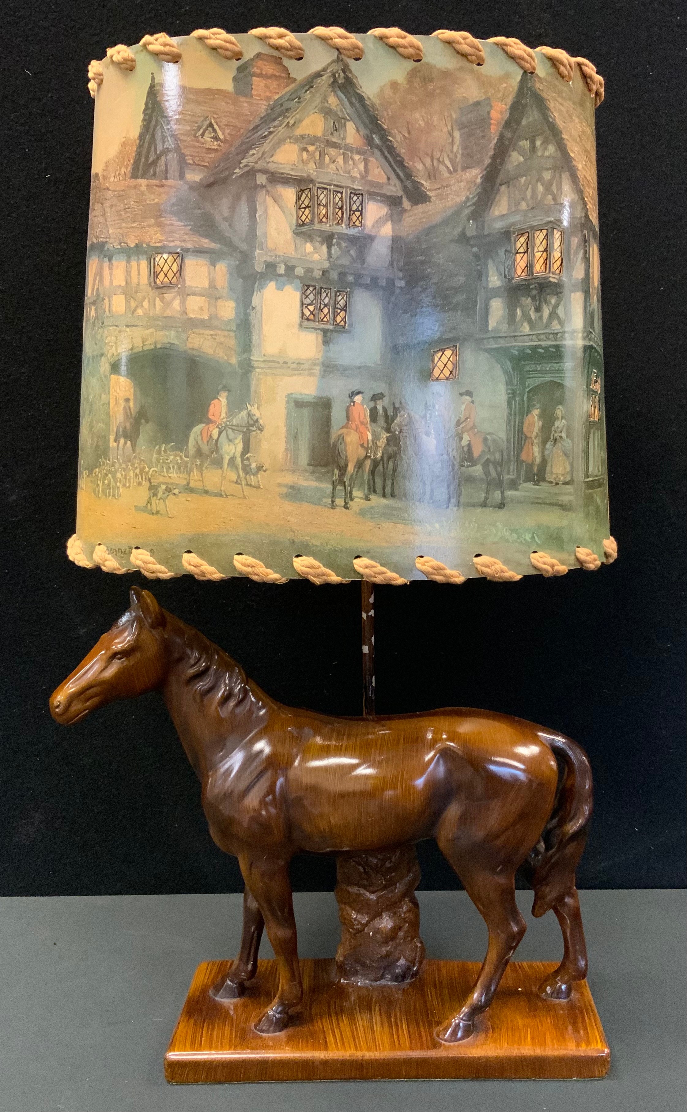 A novelty table lamp, the base with a horse, the shade with figures on horses outside a Tudor