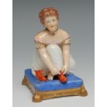 A Bloor Derby figure, of a young child putting on her boots, on a tasselled blue cushion, 9cm