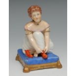 A Bloor Derby figure, of a young child putting on her boots, on a tasselled blue cushion, 9cm