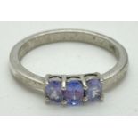 A white metal trilogy dress ring set with 3 oval cut tanzanite stones. Inside of band marked P B and