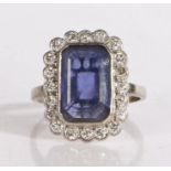 A white metal sapphire and diamond ring, the emerald cut sapphire is bezel set, surrounded by a