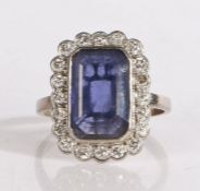 A white metal sapphire and diamond ring, the emerald cut sapphire is bezel set, surrounded by a