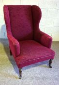 A late Victorian wing armchair, currently upholstered in claret, with turned front legs and castors