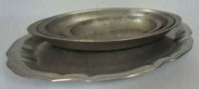 Four graduated pewter serving dishes, together with a larger scalloped tray and a brass helmet